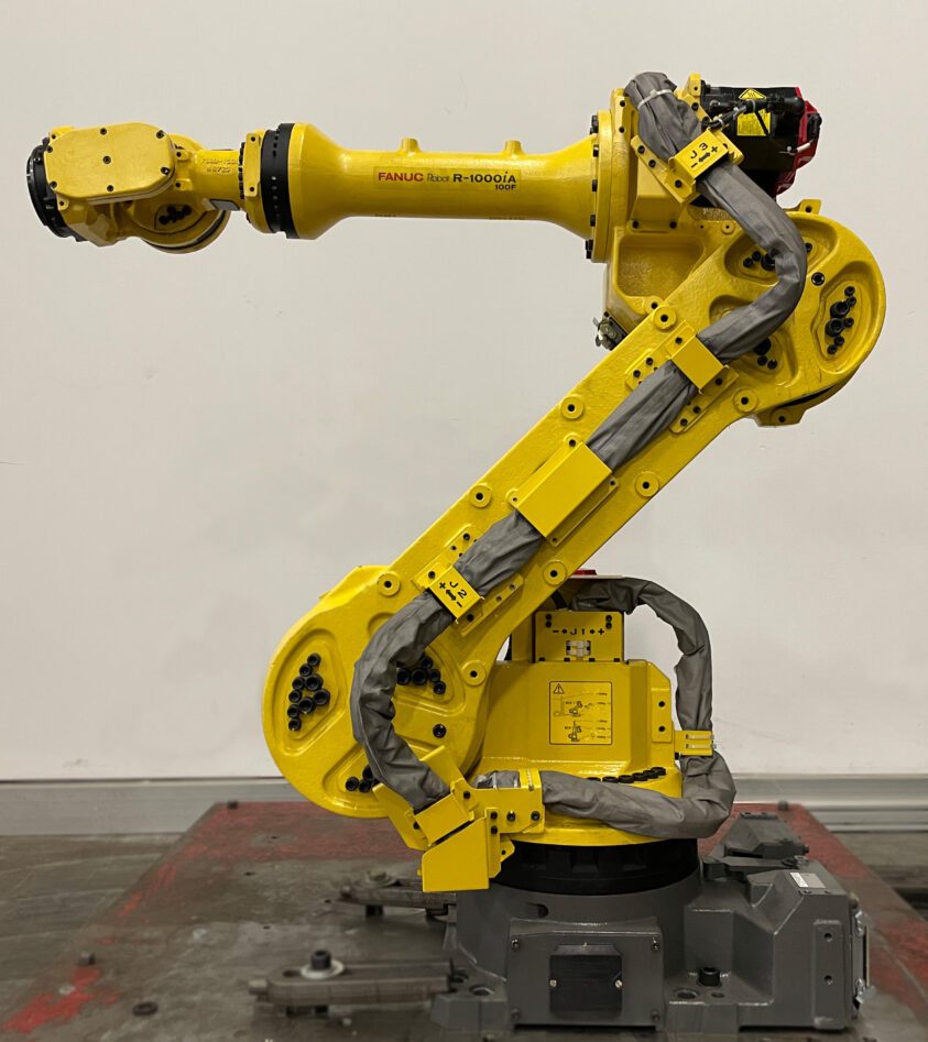 A refurbished Fanuc R-1000iA/100F R-30iB+ industrial robot in a manufacturing setting. It has a grey robotic arm with various joints and an end-effector for handling objects.